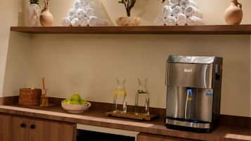 A kitchenette with a coffee maker, a bowl of apples, and a shelf with towels and plants.