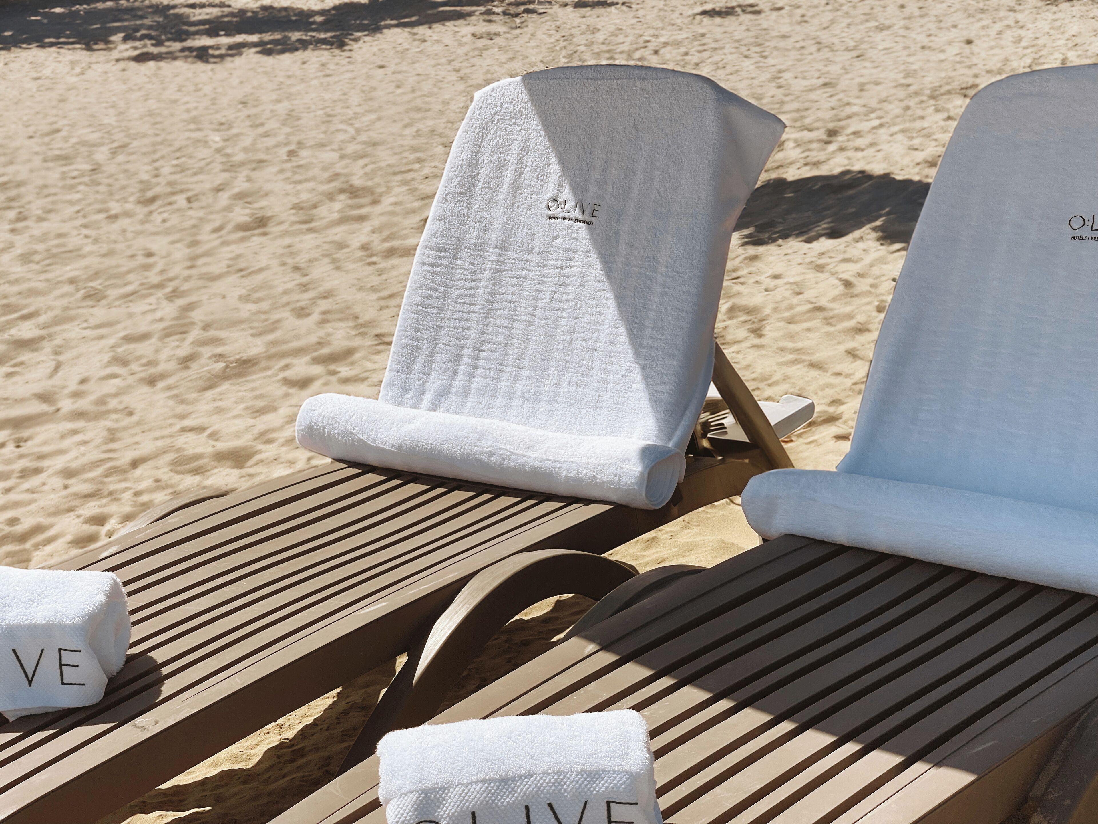 Dos sillas de playa con toallas blancas, una con la etiqueta "O:LIVE" y la otra con la etiqueta "O:LIVE HOTEL".