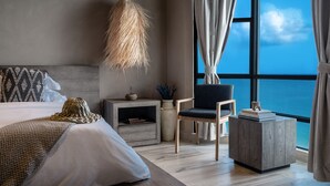 Ein modernes Schlafzimmer mit einem großen Bett, einem Nachttisch, einem Sessel und Blick aufs Meer.