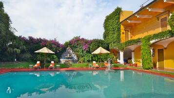 A swimming pool with lounge chairs and umbrellas, surrounded by lush greenery and a building with balconies.