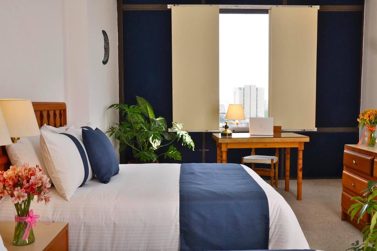 A hotel room with a bed, desk, chair, dresser, and a potted plant.