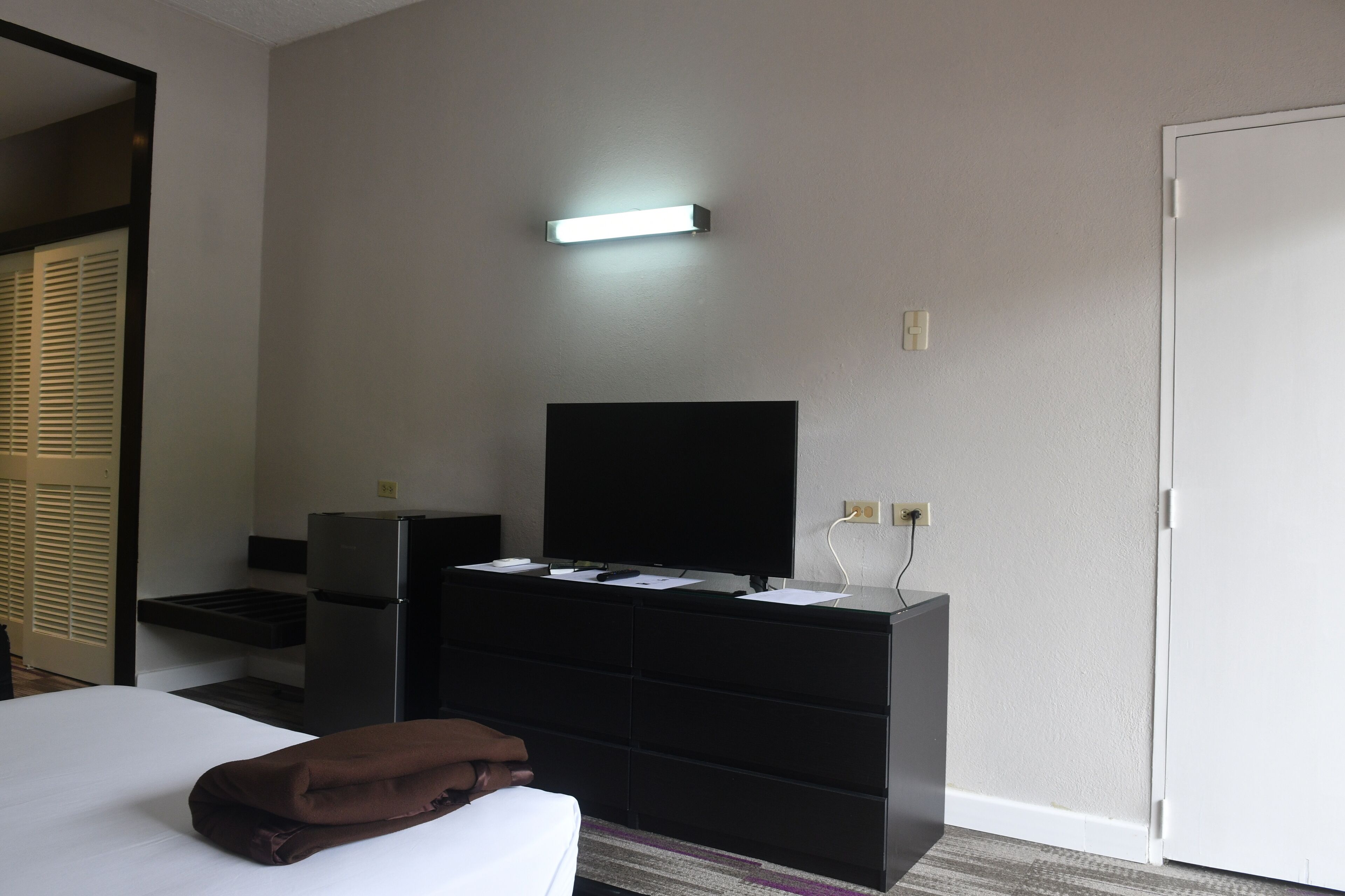 Une chambre d’hôtel avec un lit, une commode noire dotée d’un téléviseur à écran plat, un petit bureau et une porte.