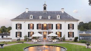 Un grand bâtiment blanc à la façade symétrique, doté de nombreuses fenêtres avec volets et d’une entrée centrale.