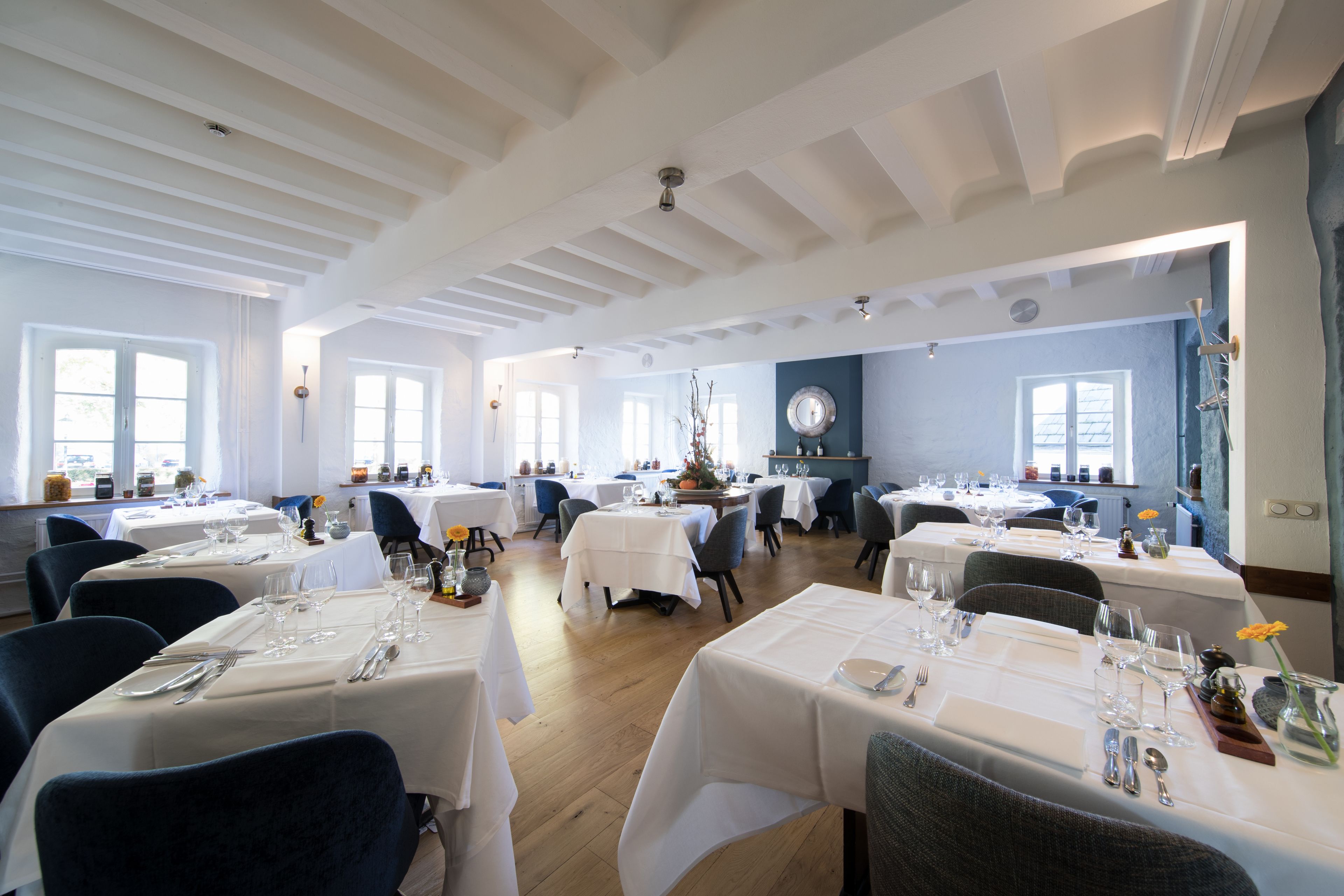 A spacious dining area with multiple tables set for dining, featuring white tablecloths and a variety of chairs.