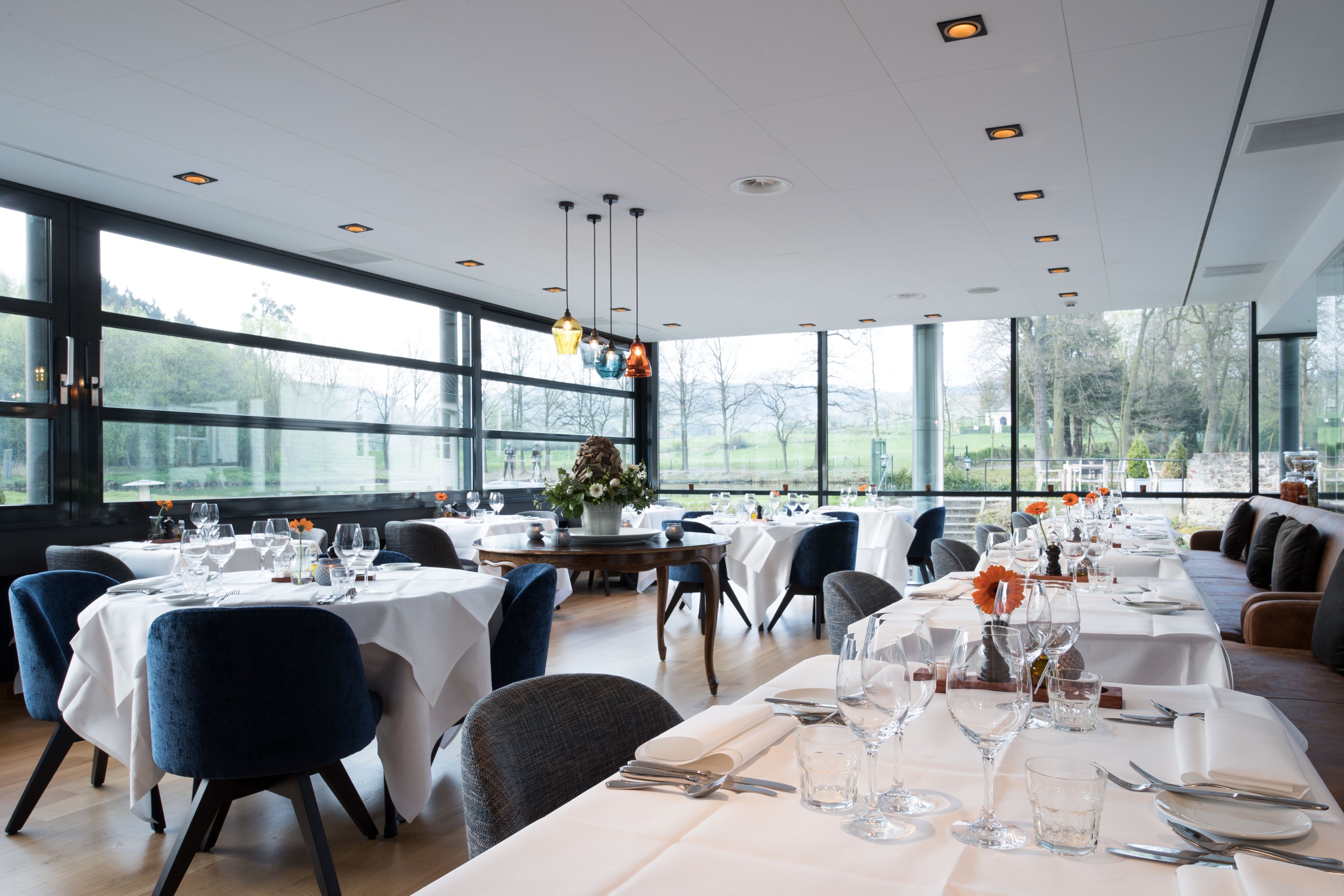 A restaurant with multiple tables set for dining, large windows offering a view of the outdoors, and modern lighting fixtures.