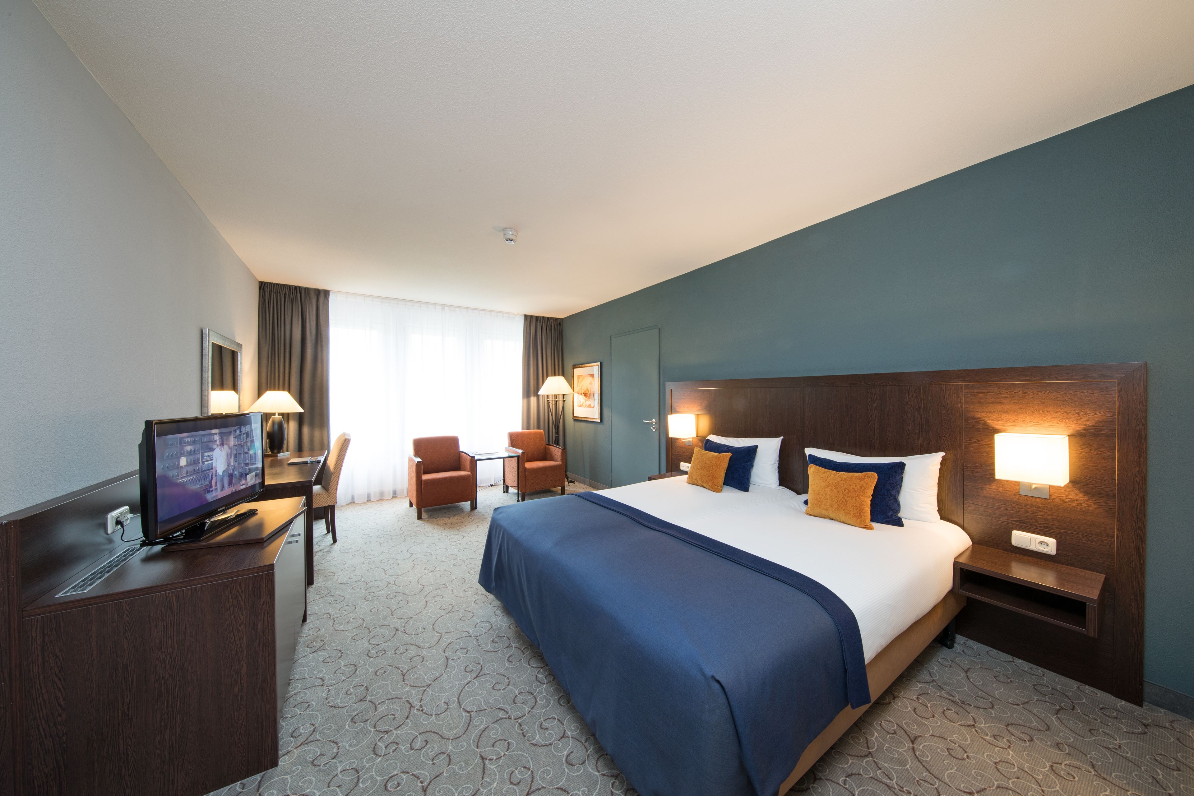Ein Hotelzimmer mit einem großen Bett, einem Schreibtisch mit Computer, zwei Stühlen und einem Fernseher.