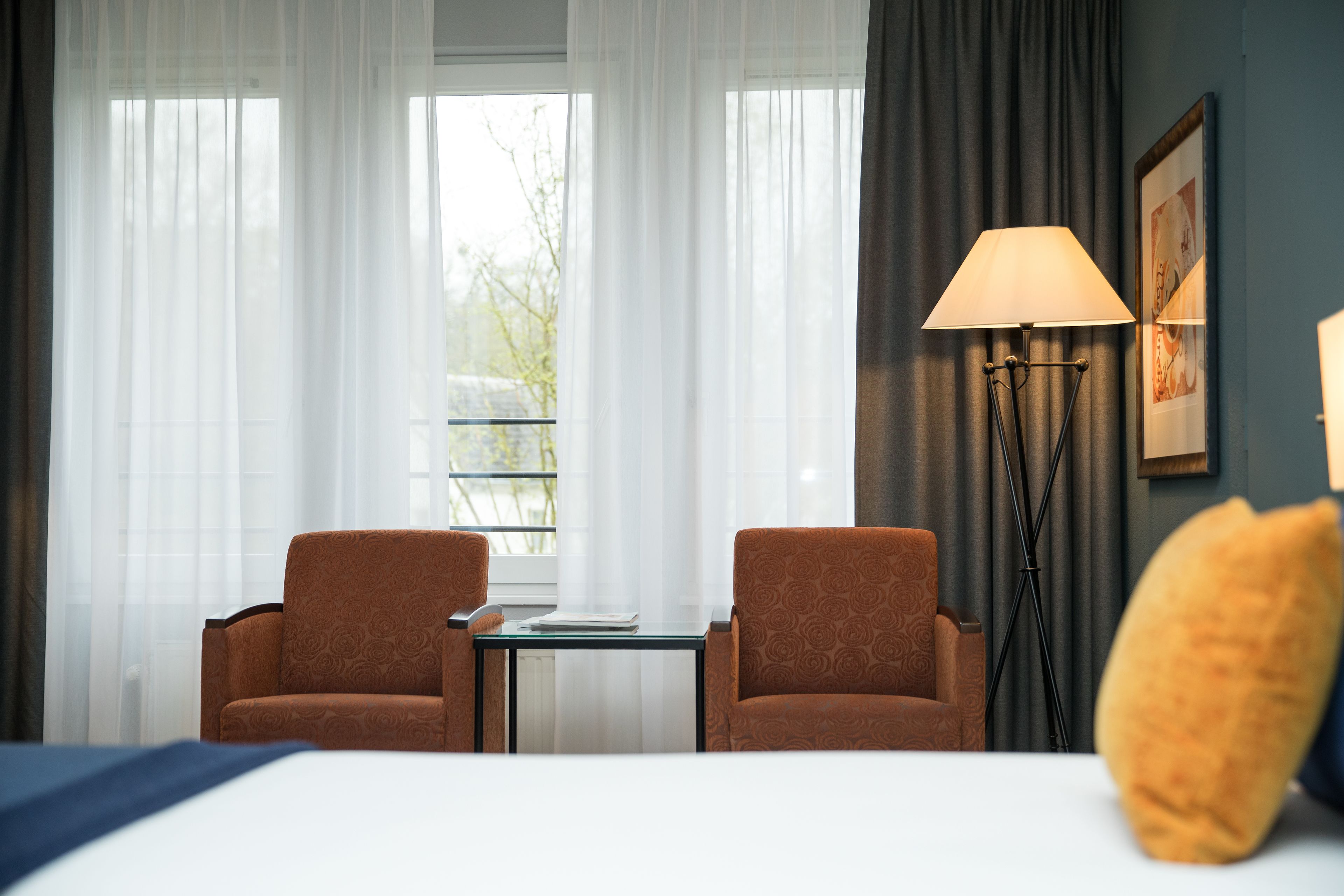 A hotel room with two brown armchairs, a small table, a lamp, and a bed.