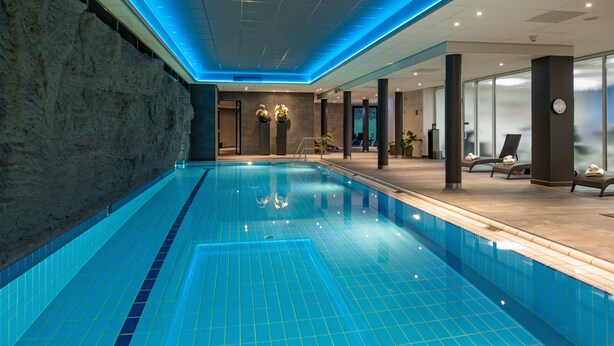 Une piscine intérieure moderne, avec des carreaux bleus, un mur en pierre et des chaises longues.