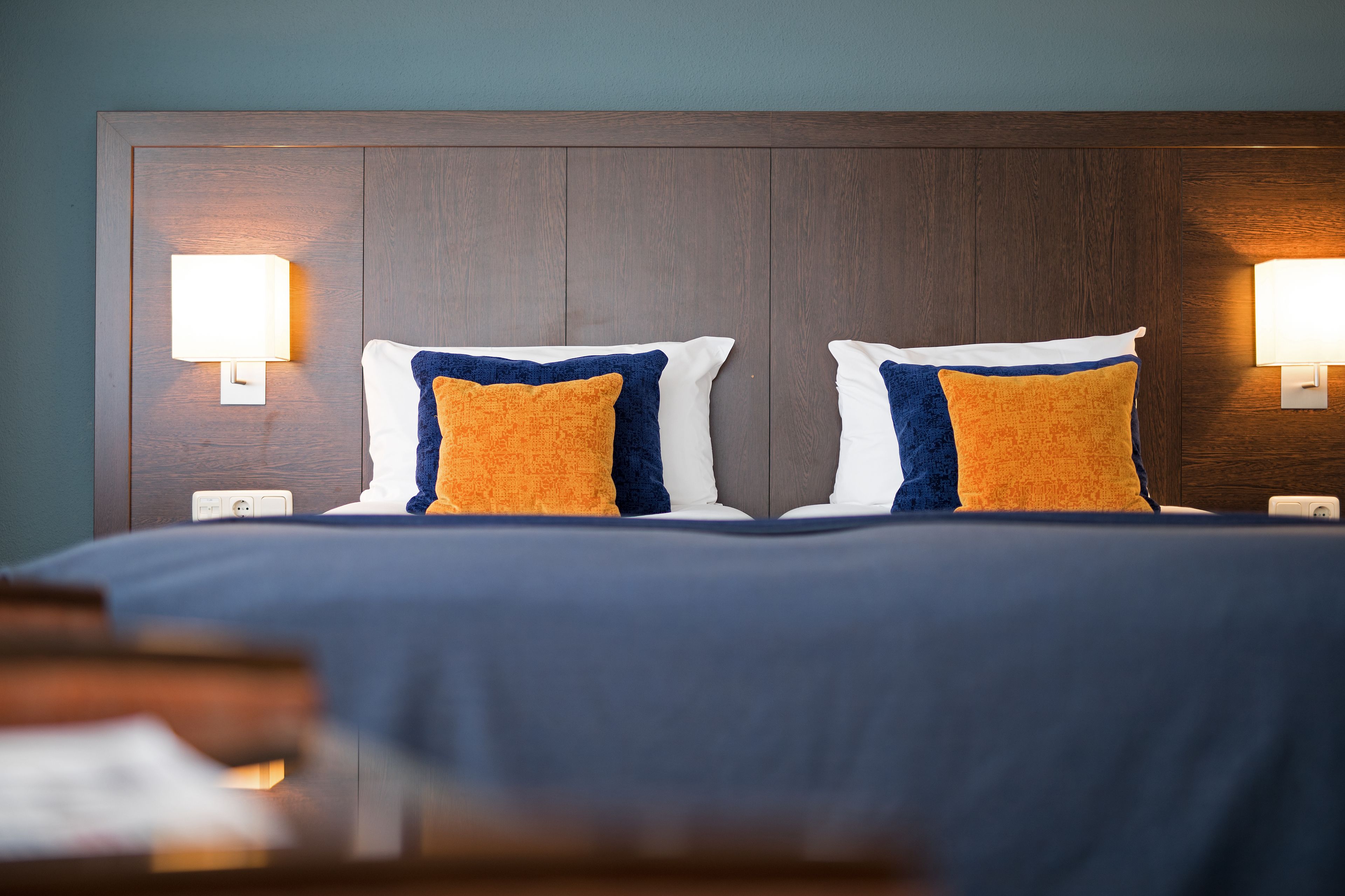Habitación de hotel con cabecera de madera, dos lámparas de noche y dos almohadas con diseño azul y naranja.