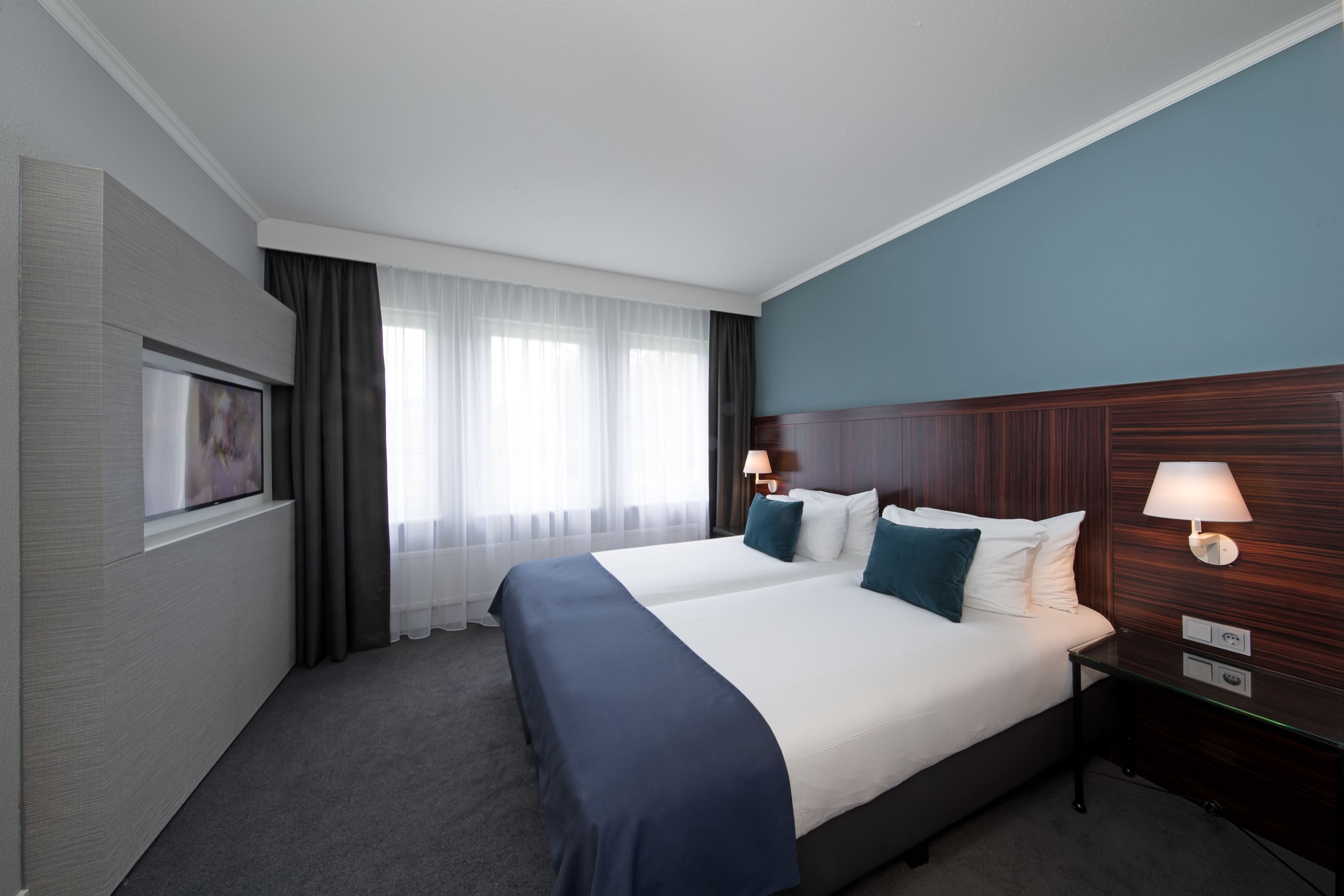 A hotel room with a large bed, two bedside tables with lamps, a flat-screen TV mounted on the wall, and a window with sheer curtains.
