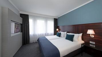 Een hotelkamer met een groot bed, twee nachtkastjes met lampen, een flatscreen televisie aan de muur en een raam met dunne gordijnen.