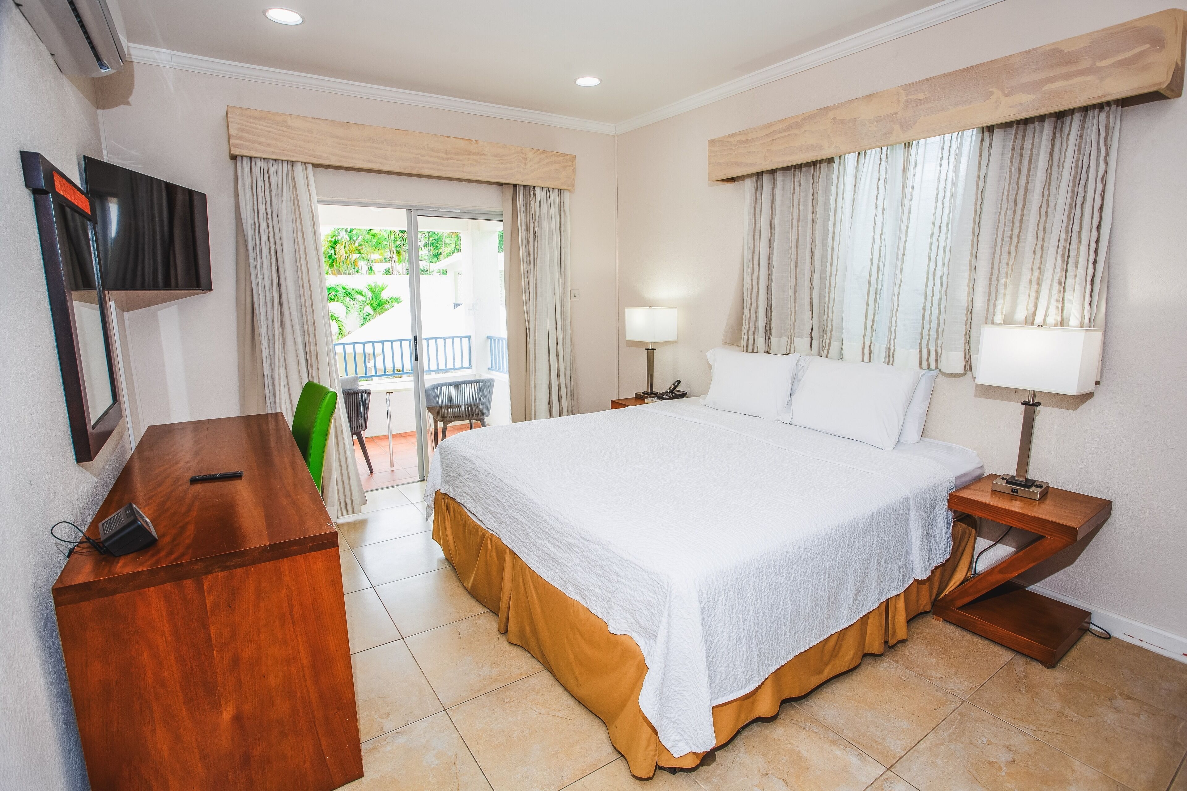 Habitación de hotel con una cama grande, un escritorio de madera, un televisor y un balcón con cortinas.