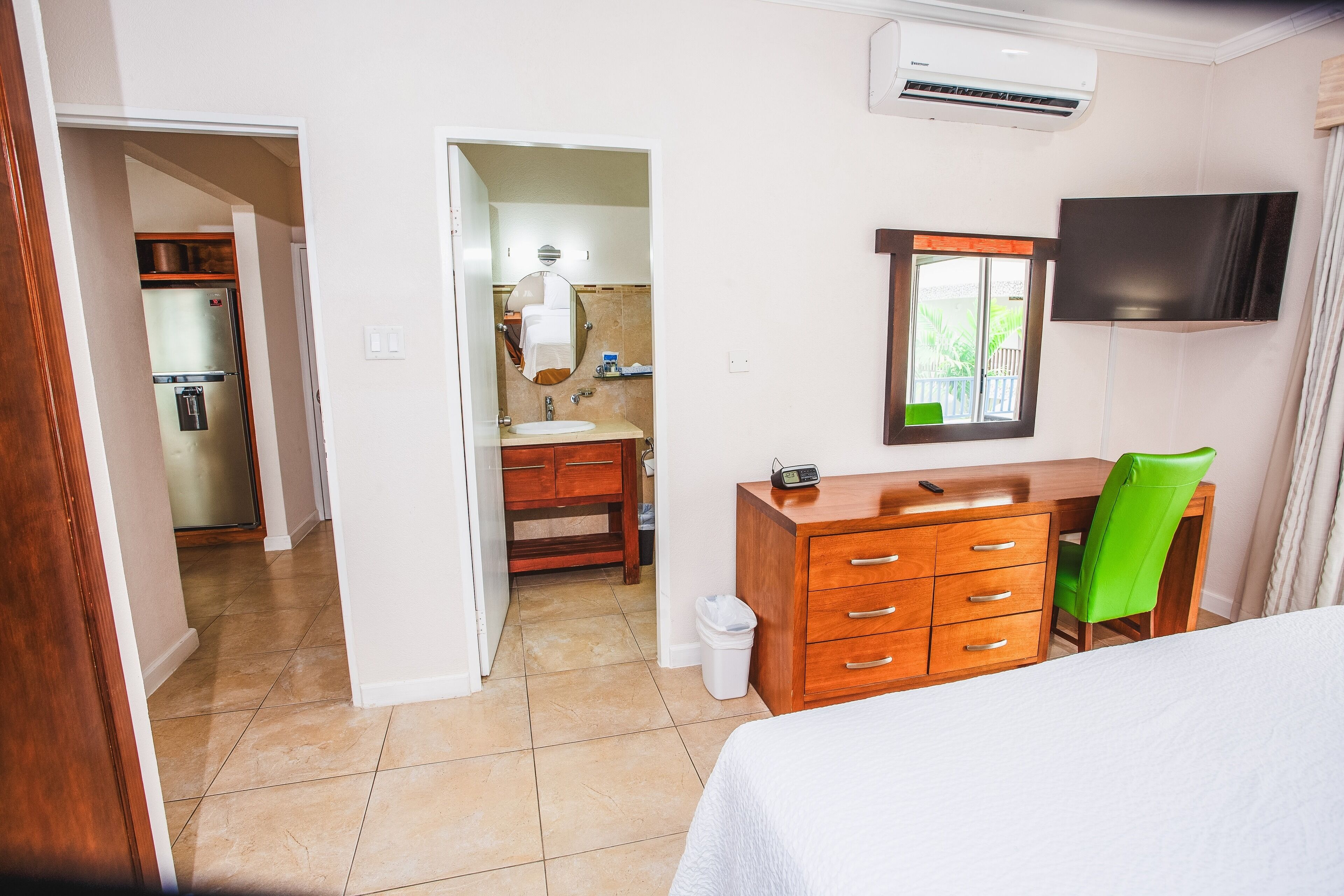 Habitació d'hotel amb un llit, un taulell de fusta, una cadira verda, una televisió i una zona de cuina visible a través d'una porta oberta.