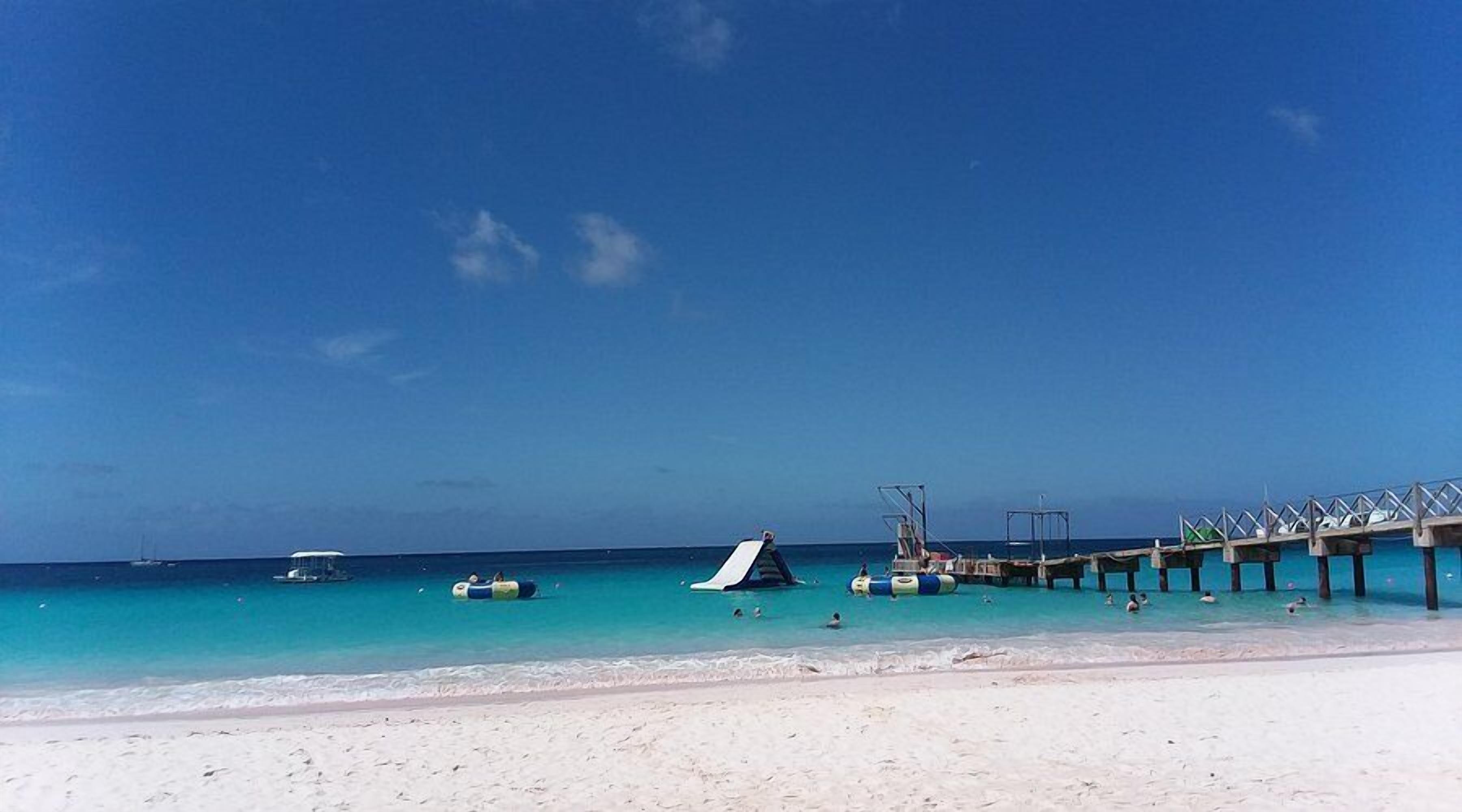 Une plage avec une jetée qui s’avance dans l’océan, une eau turquoise et limpide, et une rive sablonneuse.