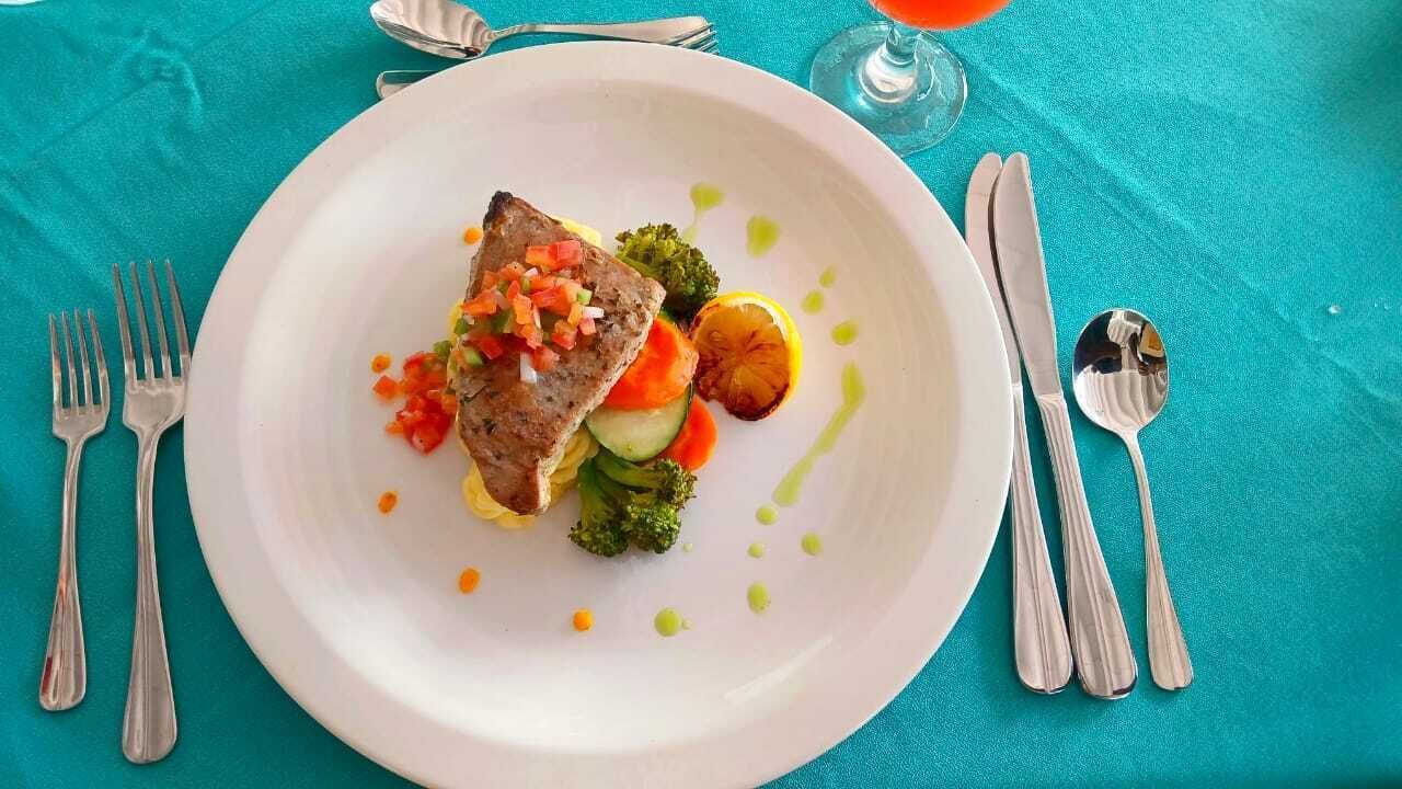 Een wit bord met vlees, groenten en saus, geplaatst op een turquoise tafelkleed met bestek.