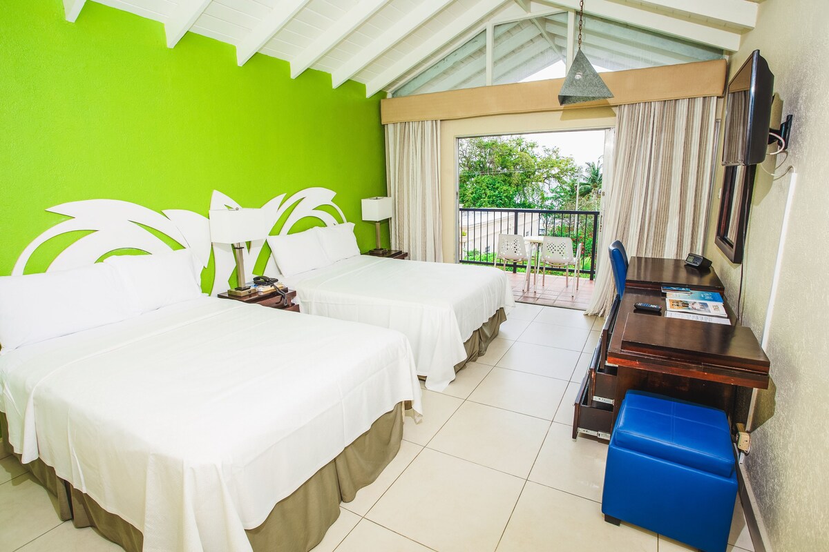 A hotel room with two beds, a desk, a chair, a TV, and a balcony with a view of greenery.