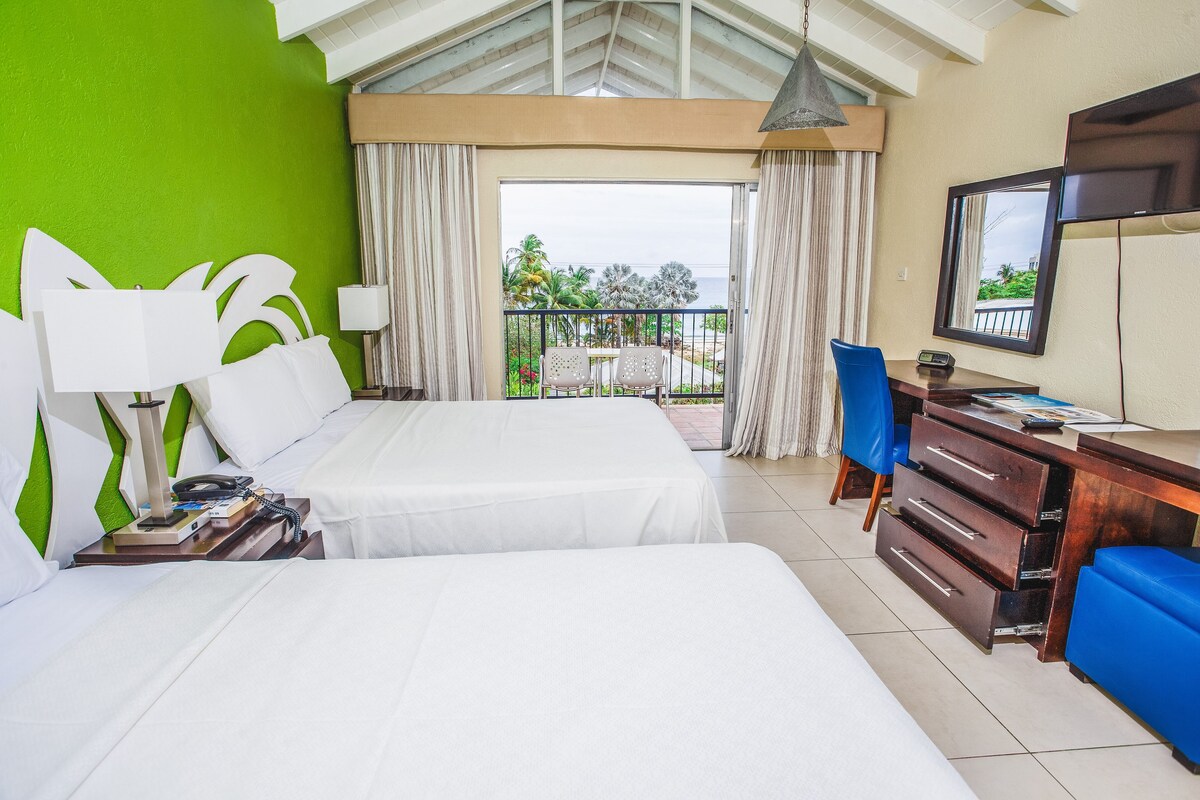 A hotel room with two beds, a desk with a chair, a TV, and a balcony with a view of the ocean.