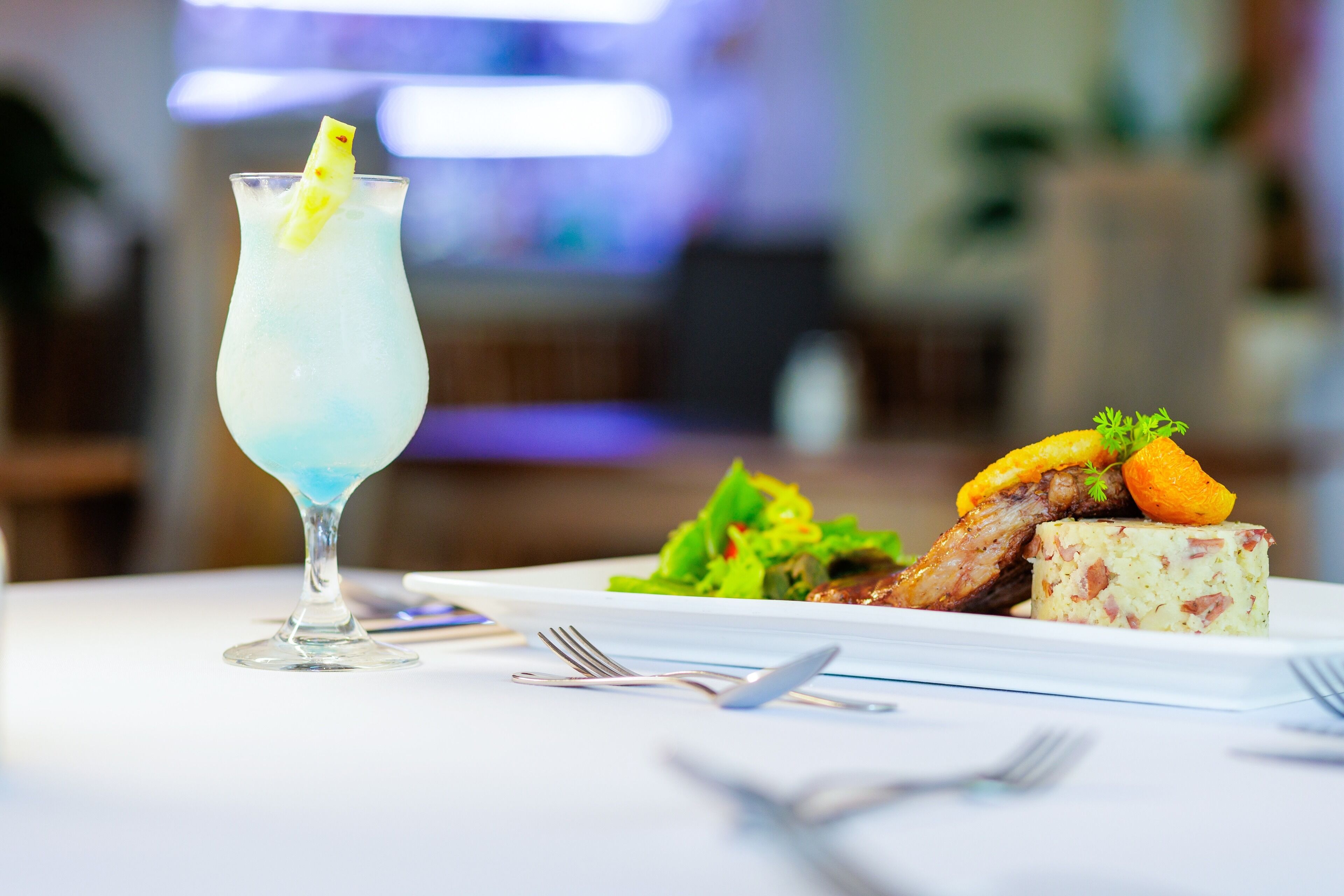 A dining table with a blue cocktail, a plate of food, and cutlery.