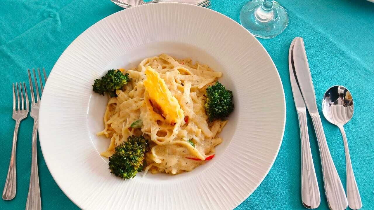 Un plato blanco con pasta, brócoli y una porción de fruta amarilla sobre un mantel turquesa.