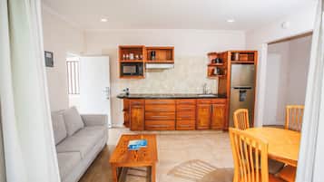A compact kitchen with wooden cabinets, a microwave, and a refrigerator. There is a dining table with chairs and a sofa in the living area.