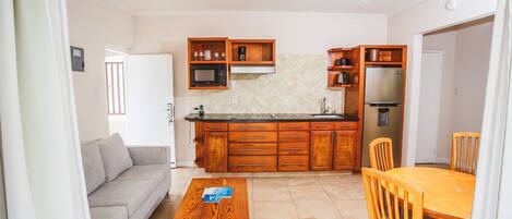 Cocina compacta con armarios de madera, microondas y refrigerador. Hay una mesa de comedor con sillas y un sofá en la sala.