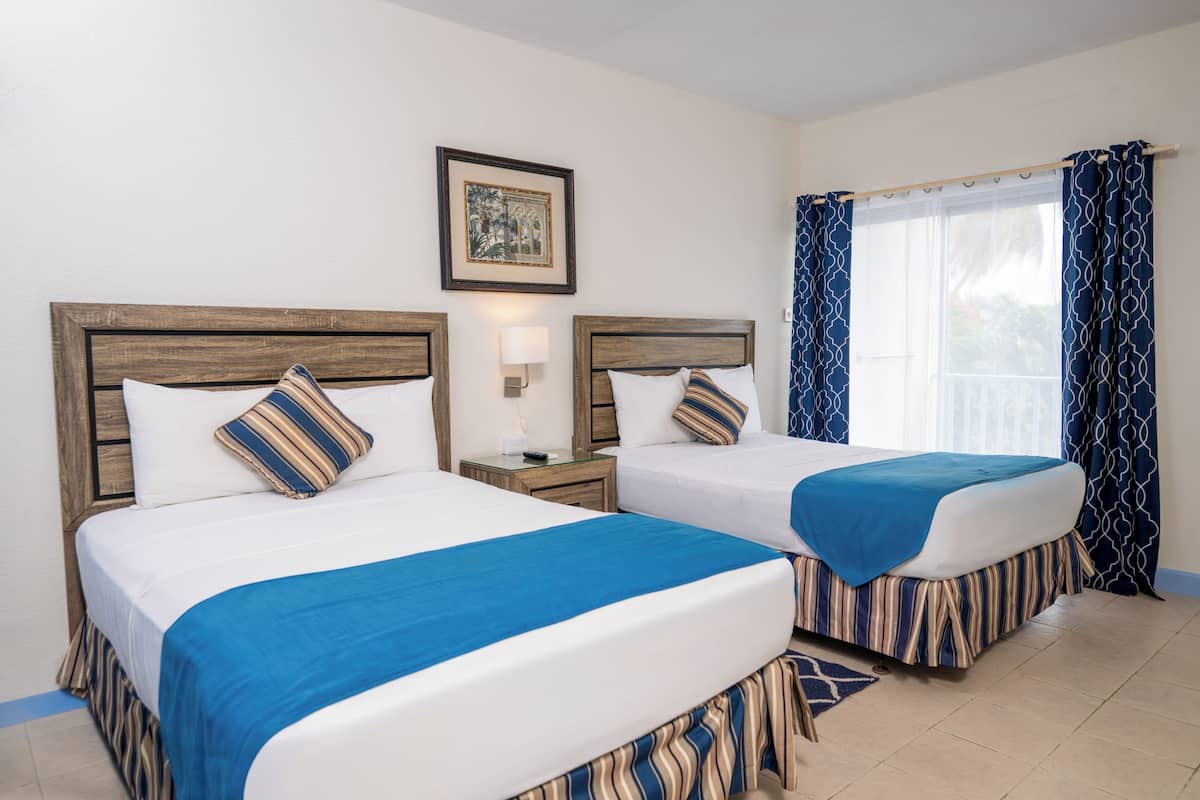 Two beds with blue and striped bedding, a nightstand with a lamp, a framed picture on the wall, and a window with blue curtains.