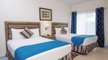 Dos camas con ropa de cama azul a rayas, una mesita de noche con lámpara, un cuadro enmarcado en la pared y una ventana con cortinas azules.