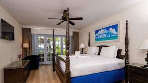 Un lit à baldaquin avec un ventilateur de plafond, un tableau représentant une plage et une vue à travers les portes vitrées.