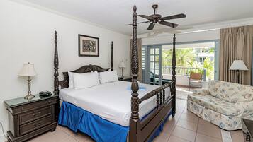 A four-poster bed with a ceiling fan, a nightstand with a lamp, a chair with a patterned cushion, and a balcony with a view of greenery.