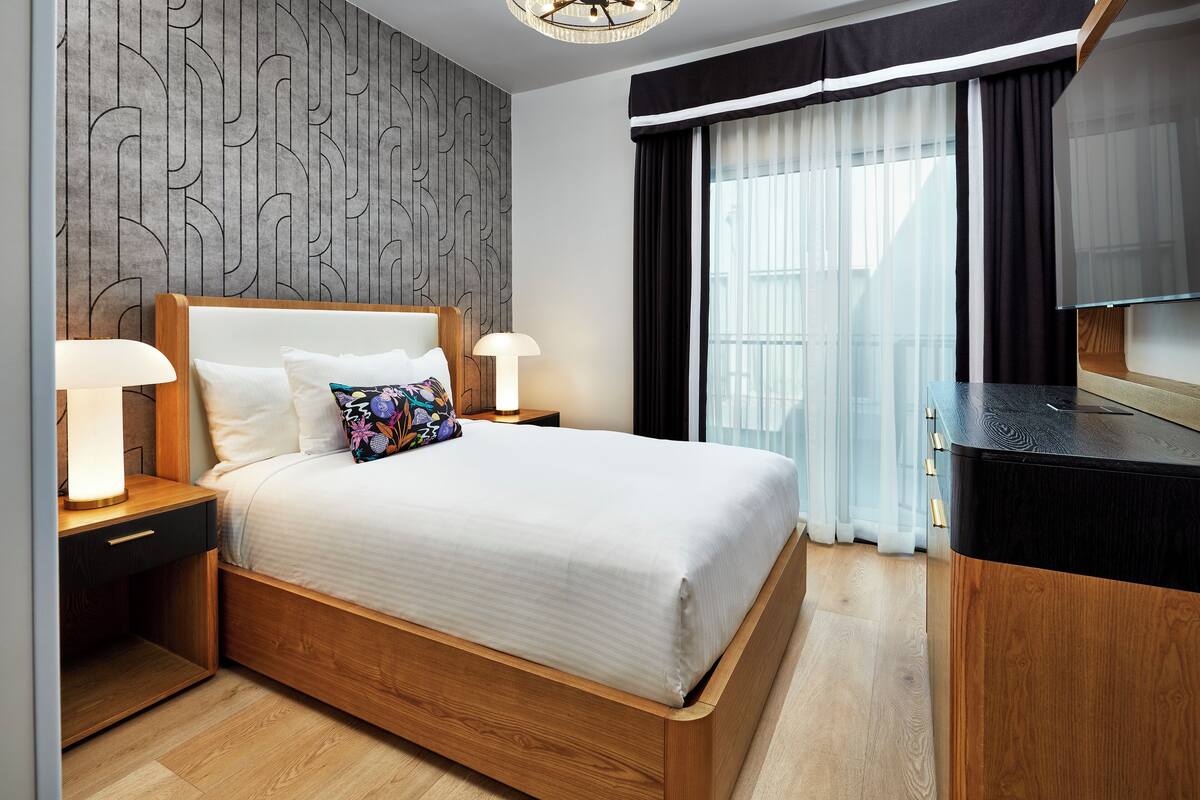 A modern hotel room with a wooden bed, two bedside tables with lamps, a large window with sheer curtains, and a built-in TV.