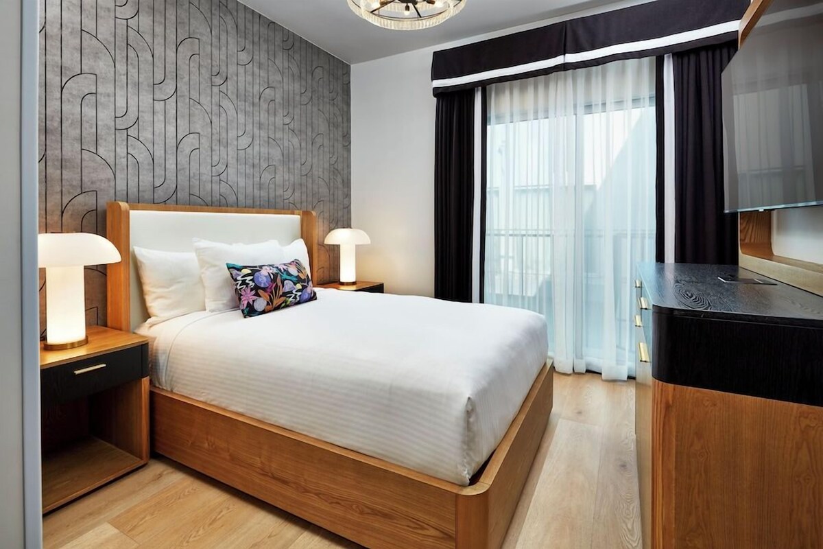 A modern hotel room with a large bed, bedside lamps, a wooden headboard, a nightstand, a desk with a flat-screen TV, and a window with sheer curtains.