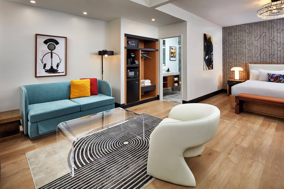 A modern hotel room with a blue sofa, a white chair, a glass coffee table, a wooden floor, a bed, a wardrobe, and a hallway leading to a bathroom.