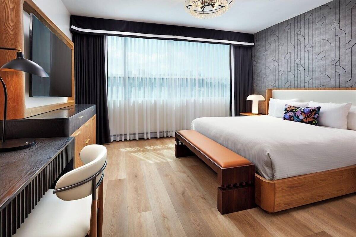 A hotel room with a bed, a desk, a chair, a bench, a TV, and a window with curtains.