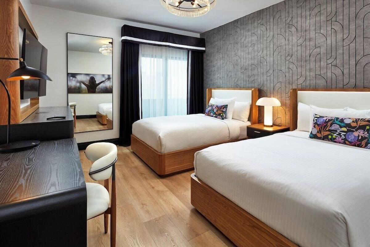 A hotel room with two beds, a desk, a chair, a TV, and a large window with curtains.