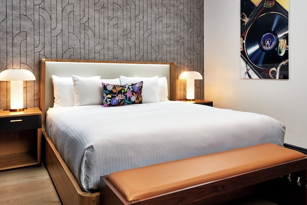 Un lit bien fait, recouvert d’une couette blanche et agrémenté d’un coussin coloré, avec deux lampes de chevet placées de part et d’autre sur des tables de nuit en bois.