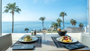 Mesa para dos con platos de pasta y verduras, dos copas de vino blanco y una cesta de pan, con vistas a una playa con palmeras y el océano.