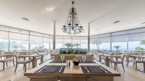 Un comedor amplio con techos altos, ventanas grandes y una lámpara central. Las mesas están preparadas con copas de vino y menús.