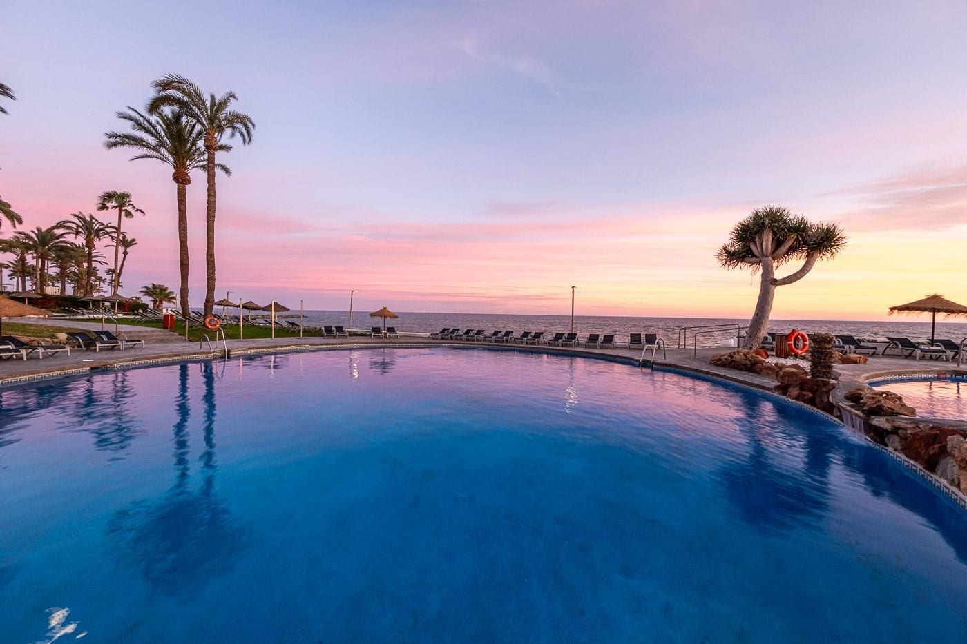 A swimming pool with lounge chairs and palm trees, surrounded by a beach and ocean view.
