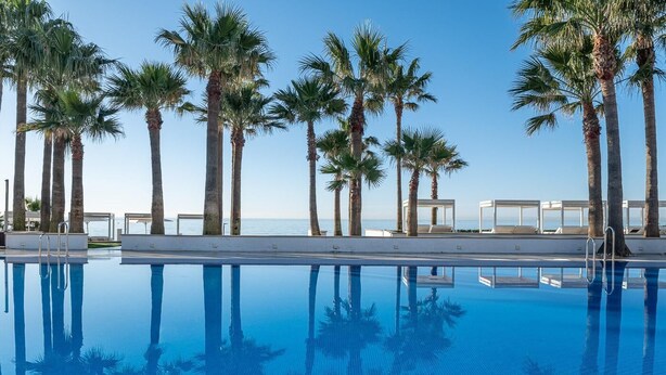 Una piscina azul cristalina con palmeras y sillas de descanso, rodeada por una zona junto a la piscina de color blanco.