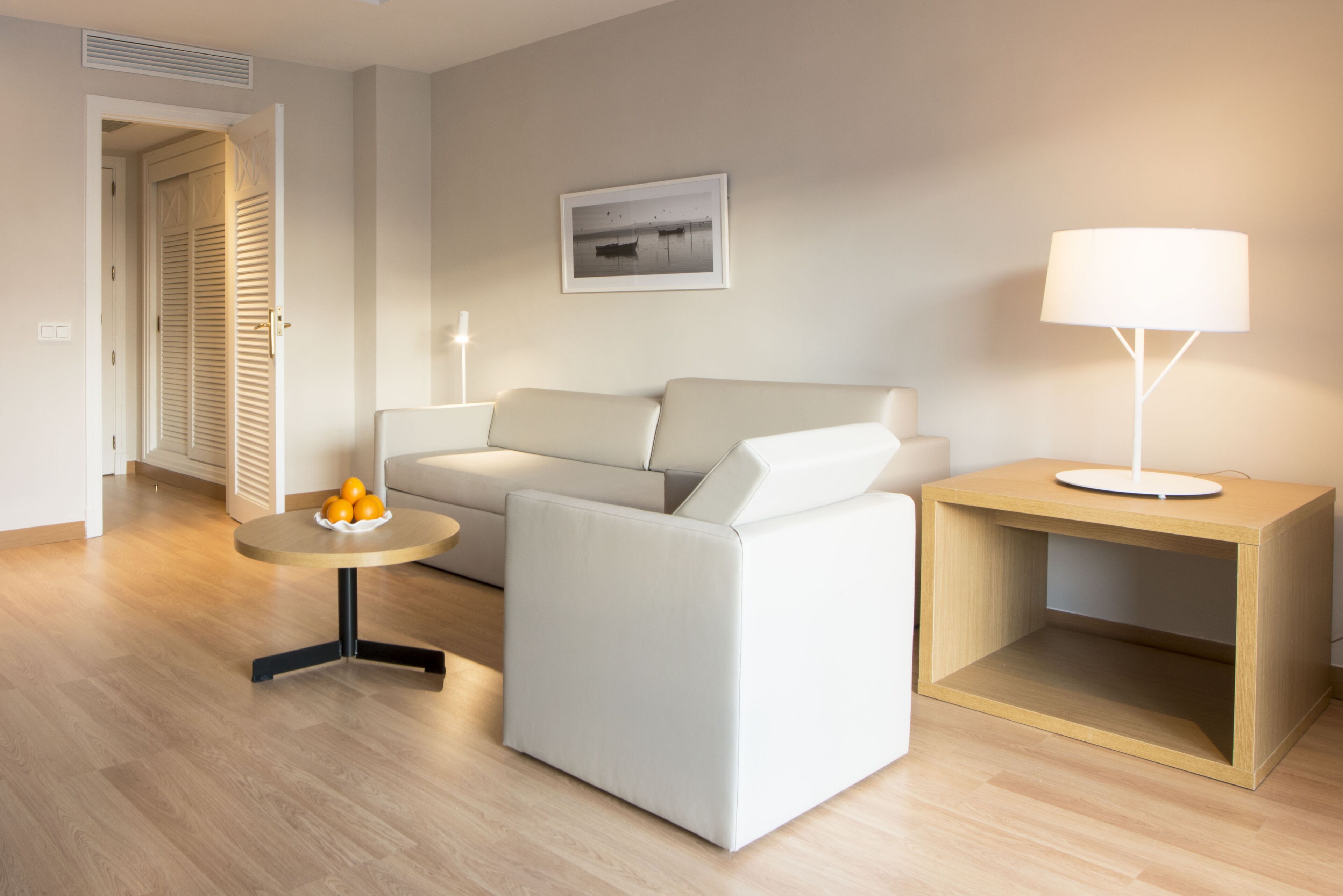 A modern living room with a sofa, a small round table with oranges, a lamp on a wooden side table, and a framed picture on the wall.