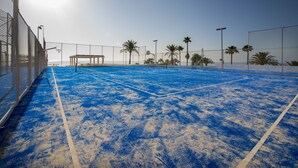 An outdoor tennis court with a blue surface, surrounded by a fence, palm trees, and a clear sky.