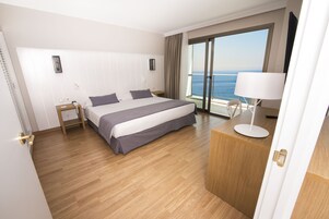 A hotel room with a large bed, a desk with a lamp, a nightstand, and a view of the sea through a large window.