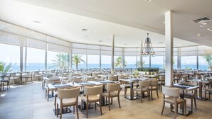 A spacious dining area with tables and chairs, large windows with a view of the ocean, and a modern chandelier.