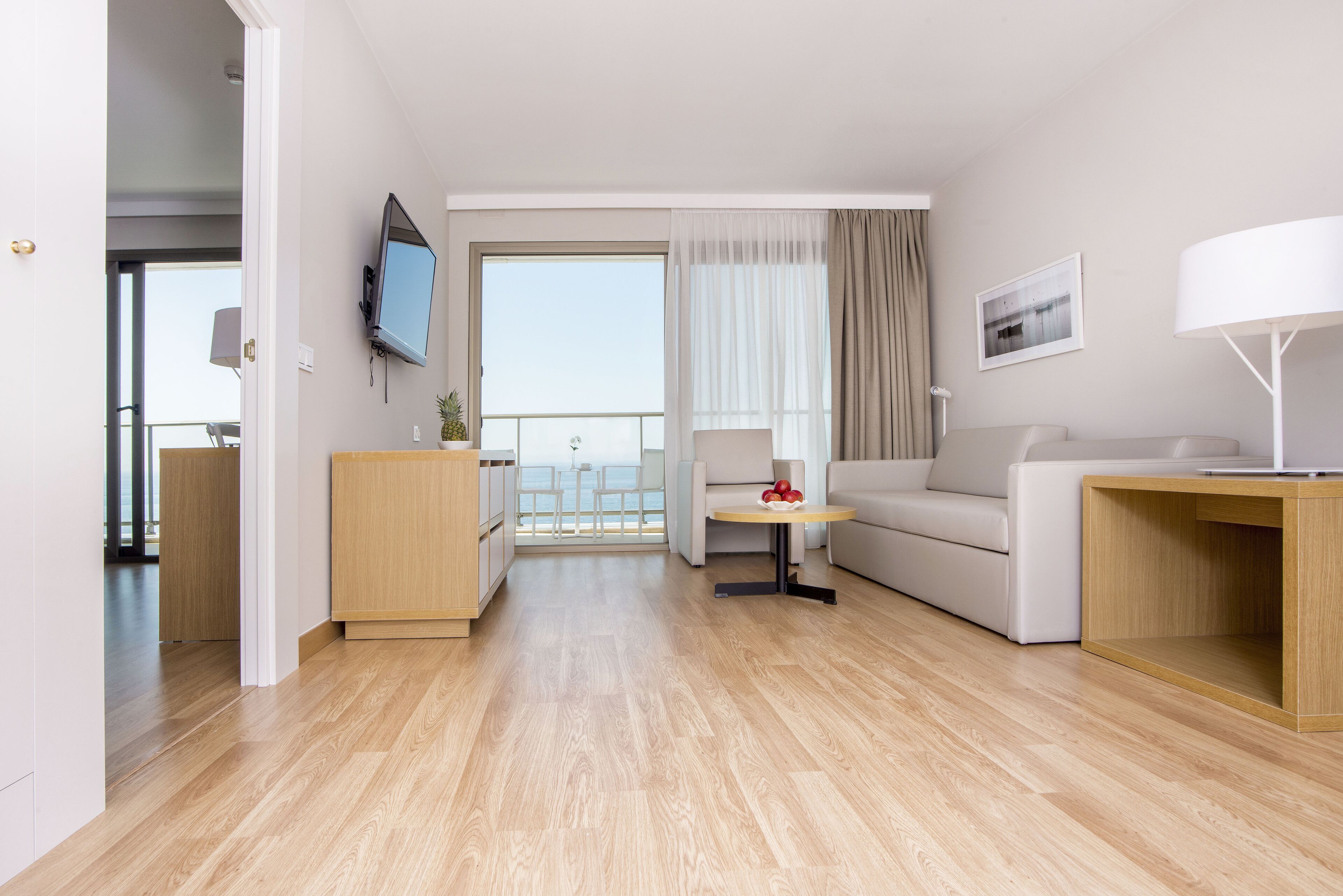 A modern hotel room with a wooden floor, a sofa, a small table with fruit, a TV, and a balcony with a view.