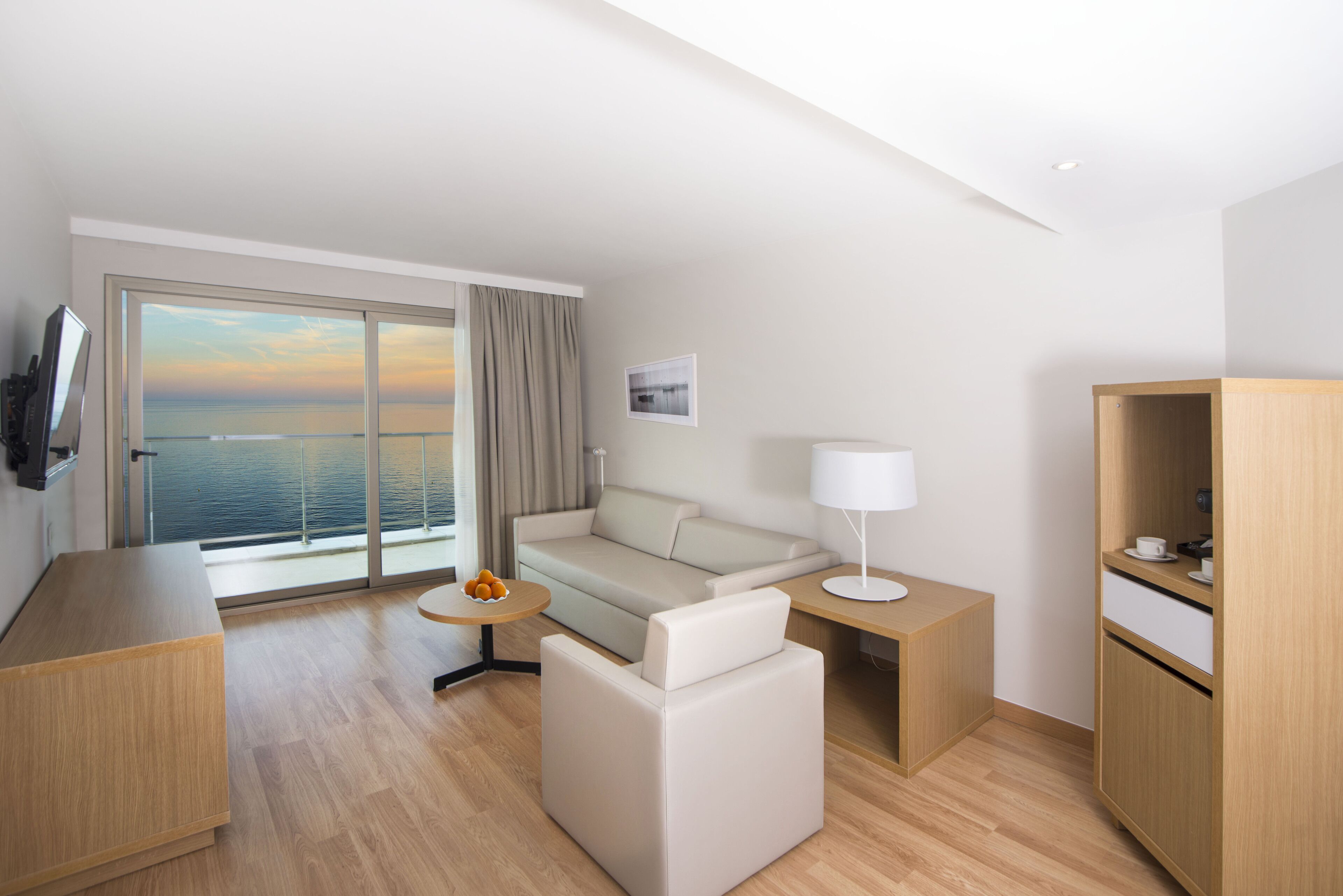 A modern hotel room with a balcony, a sofa, a small table with fruit, a lamp, and a wooden cabinet.