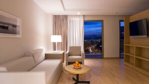 A modern hotel room with a sofa, a round table with fruit, a lamp, a TV, and a view of a cityscape at night.