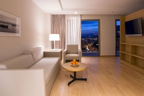 A modern hotel room with a sofa, a round table with fruit, a lamp, a TV, and a view of a cityscape at night.