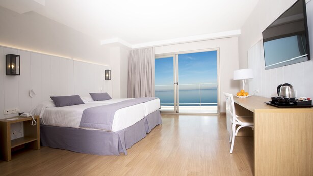 A modern hotel room with a large bed, a desk, a chair, a TV, and a view of the sea through the balcony doors.