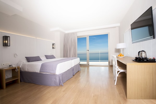 A modern hotel room with a large bed, a desk, a chair, a TV, and a view of the sea through the balcony doors.