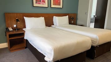 Een tweepersoonsbed met witte lakens, een houten hoofdbord, een nachtkastje met een lamp, en twee ingelijste foto's aan de muur.
